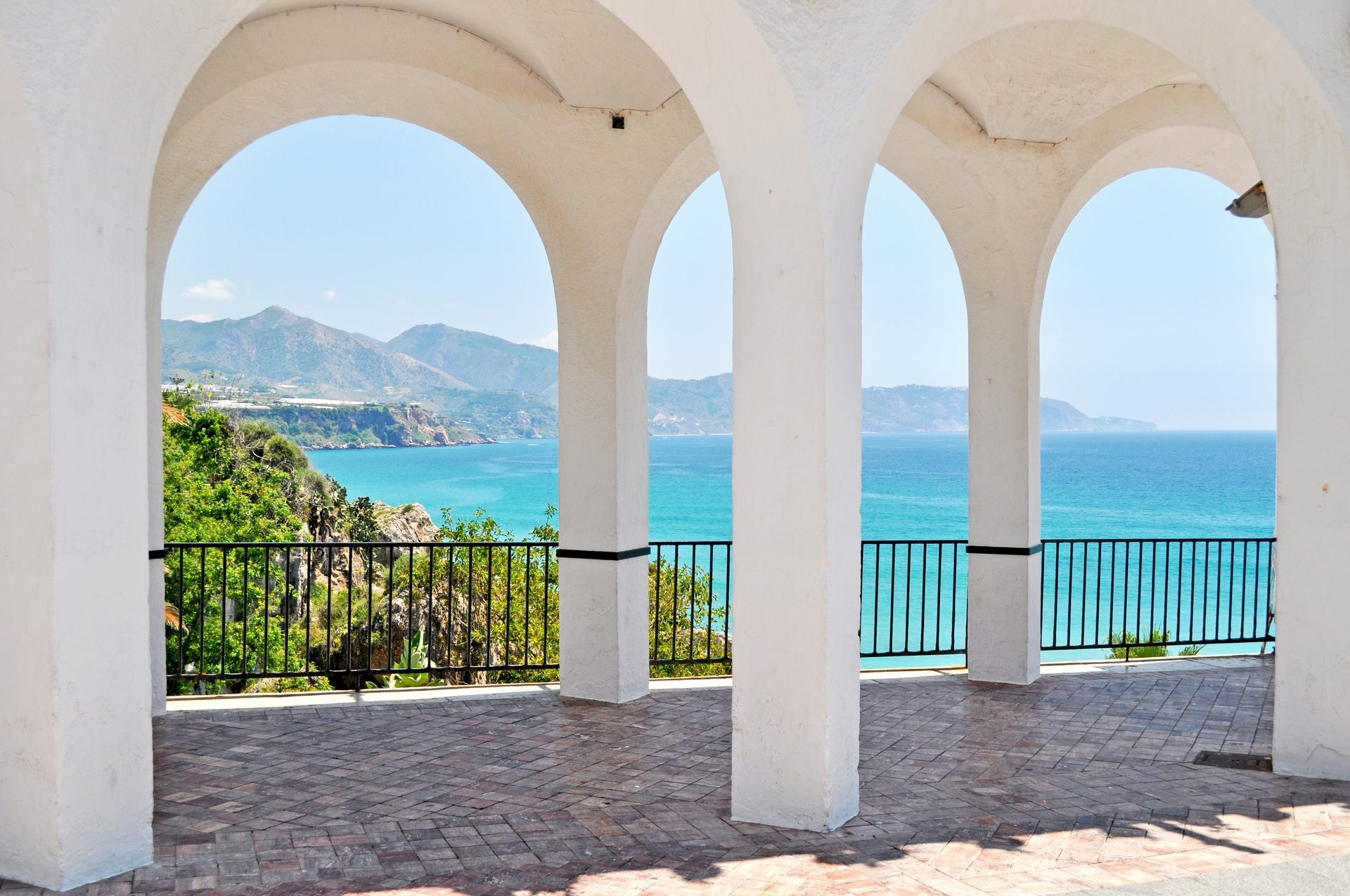 White arches balcony with ocean and mountain view, brick flooring, black railing.
